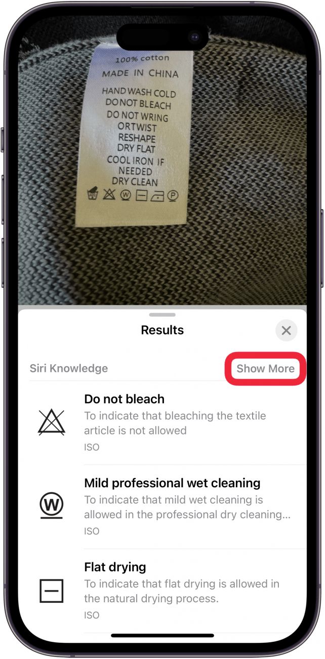 iPhone-Fotos-App, die ein Foto eines Wäscheetiketts auf einem Pullover anzeigt, wobei das visuelle Nachschlagemenü geöffnet ist und verschiedene Wäschesymbole und ihre Bedeutung anzeigt.  Um die Schaltfläche „Mehr anzeigen“ befindet sich ein rotes Kästchen